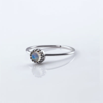 Front view of a dainty adjustable sterling silver ring with a focal AAA high quality rainbow moonstone solitaire gemstone cabochon set in a crown bezel setting. The ring is laying flat on a plain white reflective surface.