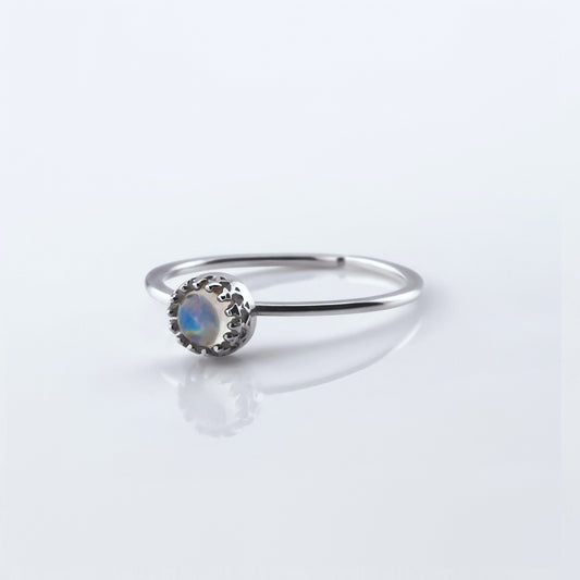 Front view of a dainty adjustable sterling silver ring with a focal AAA high quality rainbow moonstone solitaire gemstone cabochon set in a crown bezel setting. The ring is laying flat on a plain white reflective surface.