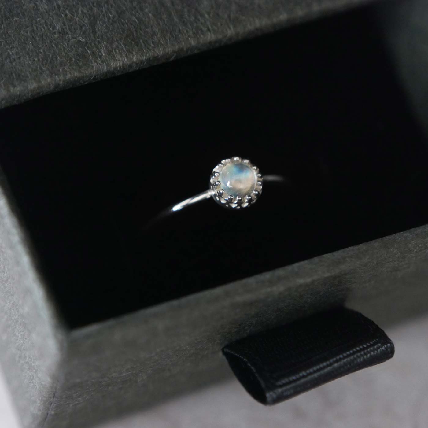 Top 3/4 view of a dainty adjustable sterling silver ring with a focal natural high quality AAA rainbow moonstone solitaire gemstone cabochon set in a crown bezel setting. The ring is tucked into the black velvet insert of a gray textured colored jewelry box with reverie blossom silver brand text on it.