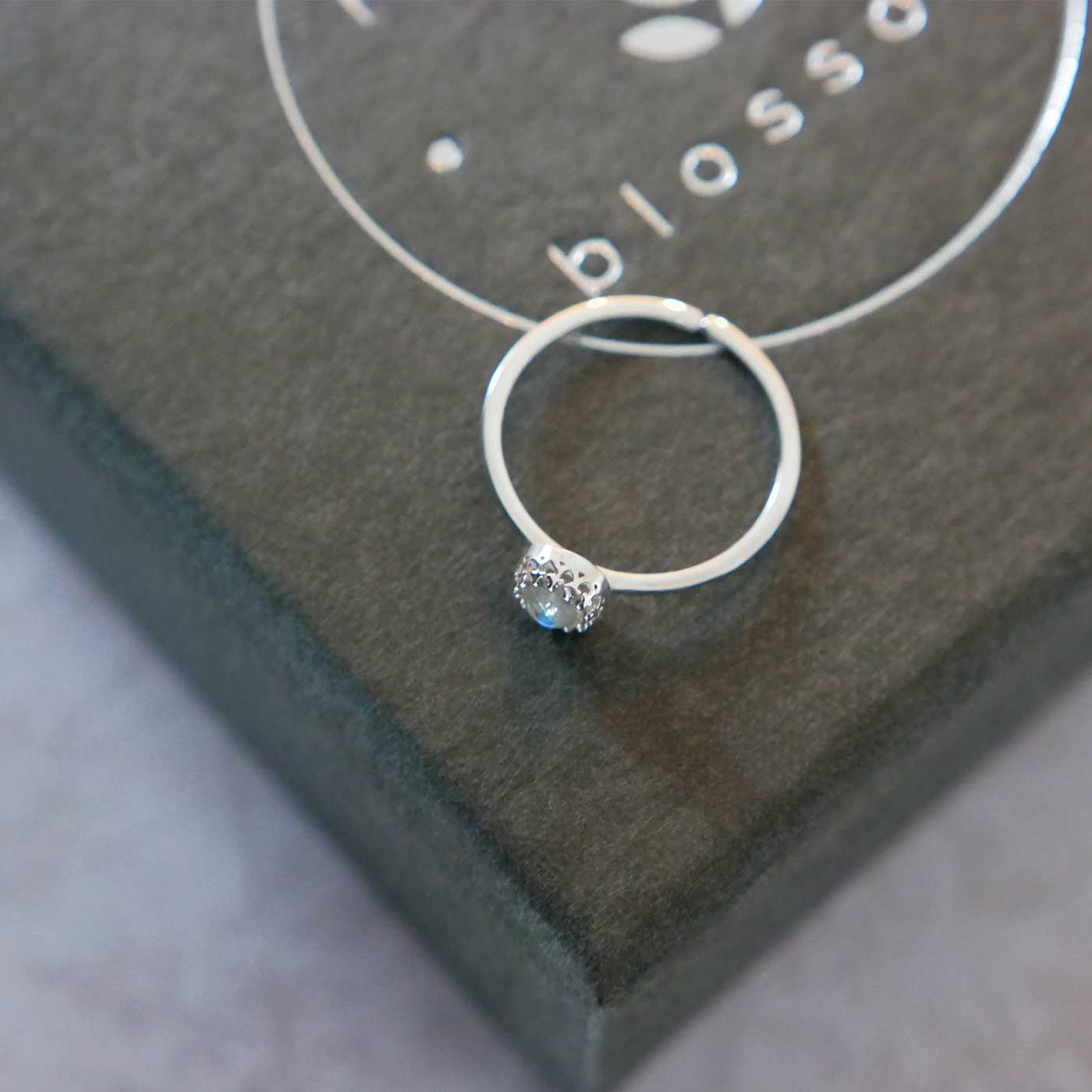 Top side view of a dainty adjustable sterling silver ring with a focal natural AAA rainbow moonstone solitaire gemstone cabochon set in a crown bezel setting. The ring is laying flat on a gray textured colored jewelry box with silver reverie blossom branded text.