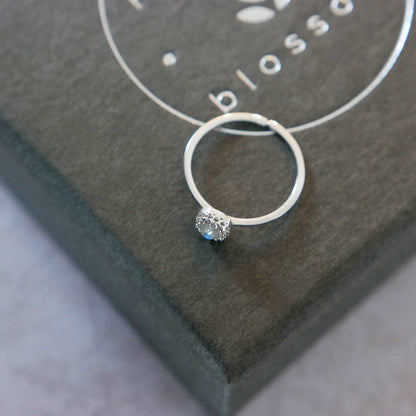 Top side view of a dainty adjustable sterling silver ring with a focal natural AAA rainbow moonstone solitaire gemstone cabochon set in a crown bezel setting. The ring is laying flat on a gray textured colored jewelry box with silver reverie blossom branded text.