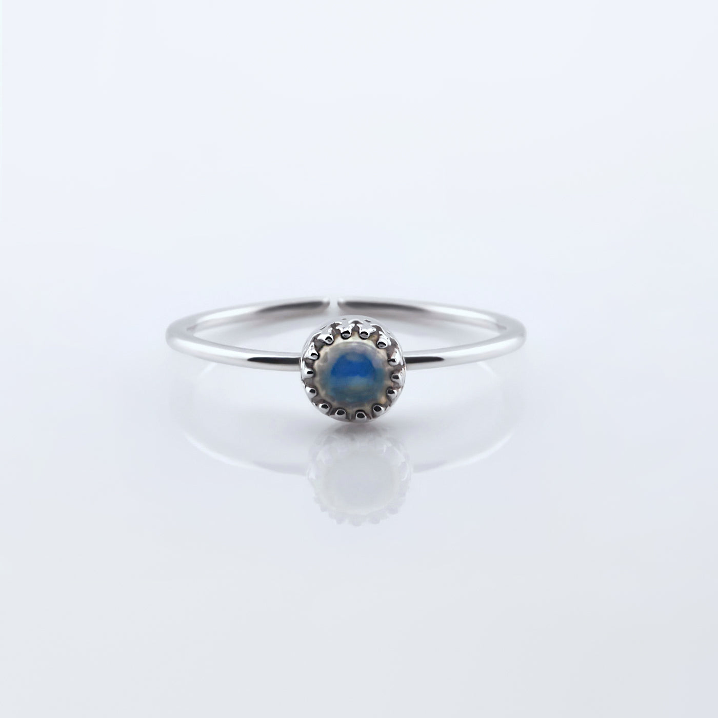 Front view of a dainty adjustable sterling silver ring with a focal high quality AAA rainbow moonstone solitaire cabochon set in a crown bezel setting. The ring is laying flat on a plain white reflective surface.