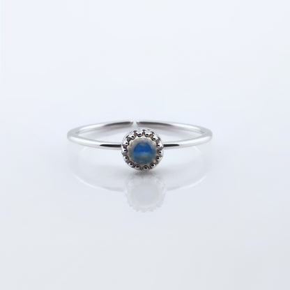 Front view of a dainty adjustable sterling silver ring with a focal high quality AAA rainbow moonstone solitaire cabochon set in a crown bezel setting. The ring is laying flat on a plain white reflective surface.