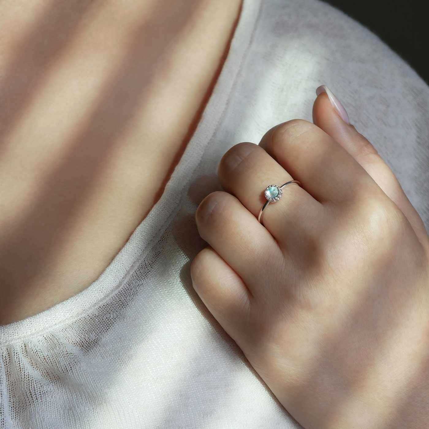 Partial view of a hand up to shoulder area of a person wearing a dainty adjustable sterling silver ring with a focal natural high quality aaa rainbow moonstone gemstone cabochon set in a crown bezel setting. The person is wearing a beige off white colored top, with window shutter shadows, and a dark gray background.