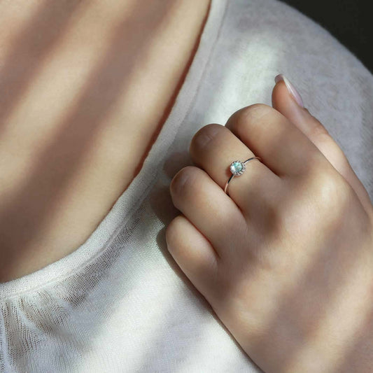 Partial view of a hand up to shoulder area of a person wearing a dainty adjustable sterling silver ring with a focal natural high quality aaa rainbow moonstone gemstone cabochon set in a crown bezel setting. The person is wearing a beige off white colored top, with window shutter shadows, and a dark gray background.