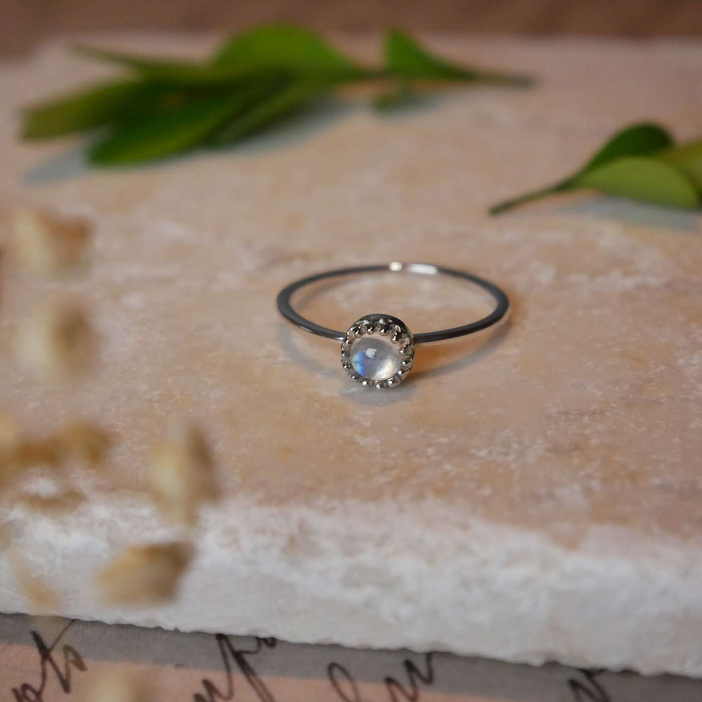 Front view of a dainty adjustable sterling silver ring with a focal natural high quality aaa rainbow moonstone solitaire gemstone cabochon set in a crown bezel setting laying on a stone surface over textured vintage beige pink paper with cursive writing on it, also stylized with green leaves and blurred beige flowers in the foreground.