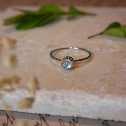 Front view of a dainty adjustable sterling silver ring with a focal natural high quality aaa rainbow moonstone solitaire gemstone cabochon set in a crown bezel setting laying on a stone surface over textured vintage beige pink paper with cursive writing on it, also stylized with green leaves and blurred beige flowers in the foreground.