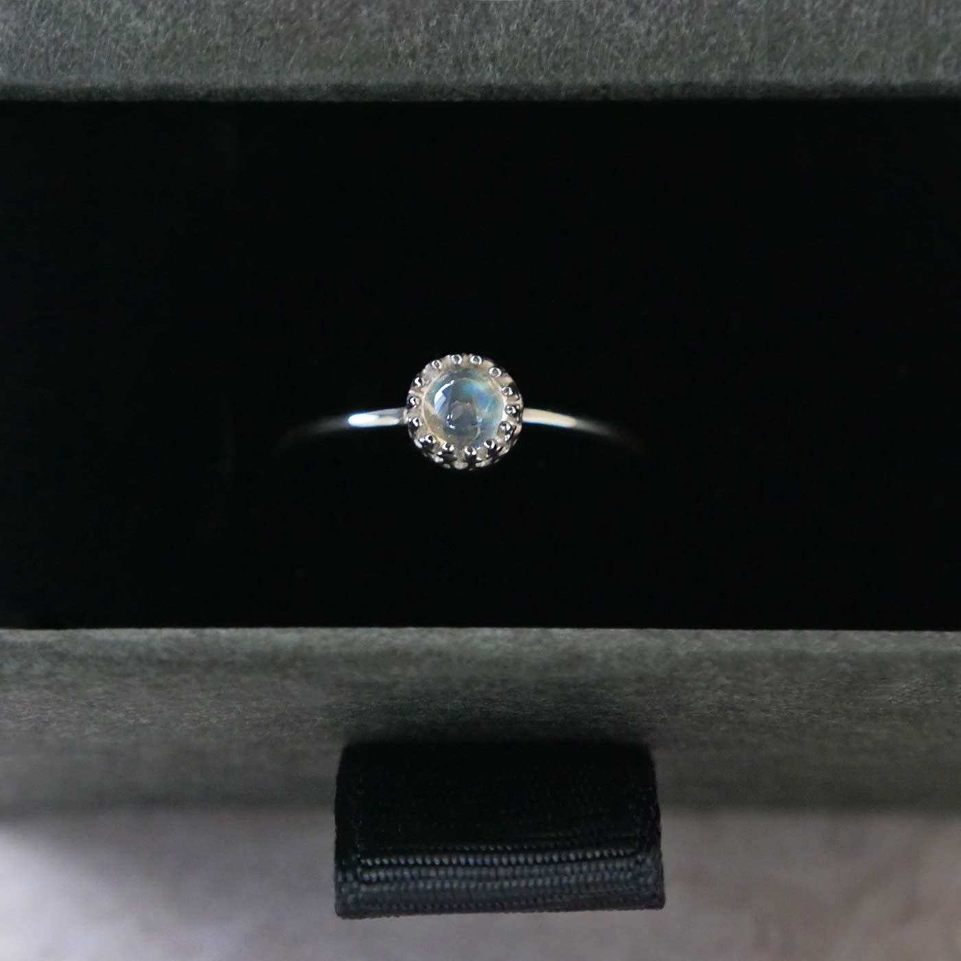 Top view of a dainty adjustable sterling silver ring with a focal natural high quality aaa rainbow moonstone solitaire gemstone cabochon set in a crown bezel setting. The ring is tucked into the black velvet insert of a gray textured colored jewelry box.