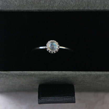 Top view of a dainty adjustable sterling silver ring with a focal natural high quality aaa rainbow moonstone solitaire gemstone cabochon set in a crown bezel setting. The ring is tucked into the black velvet insert of a gray textured colored jewelry box.