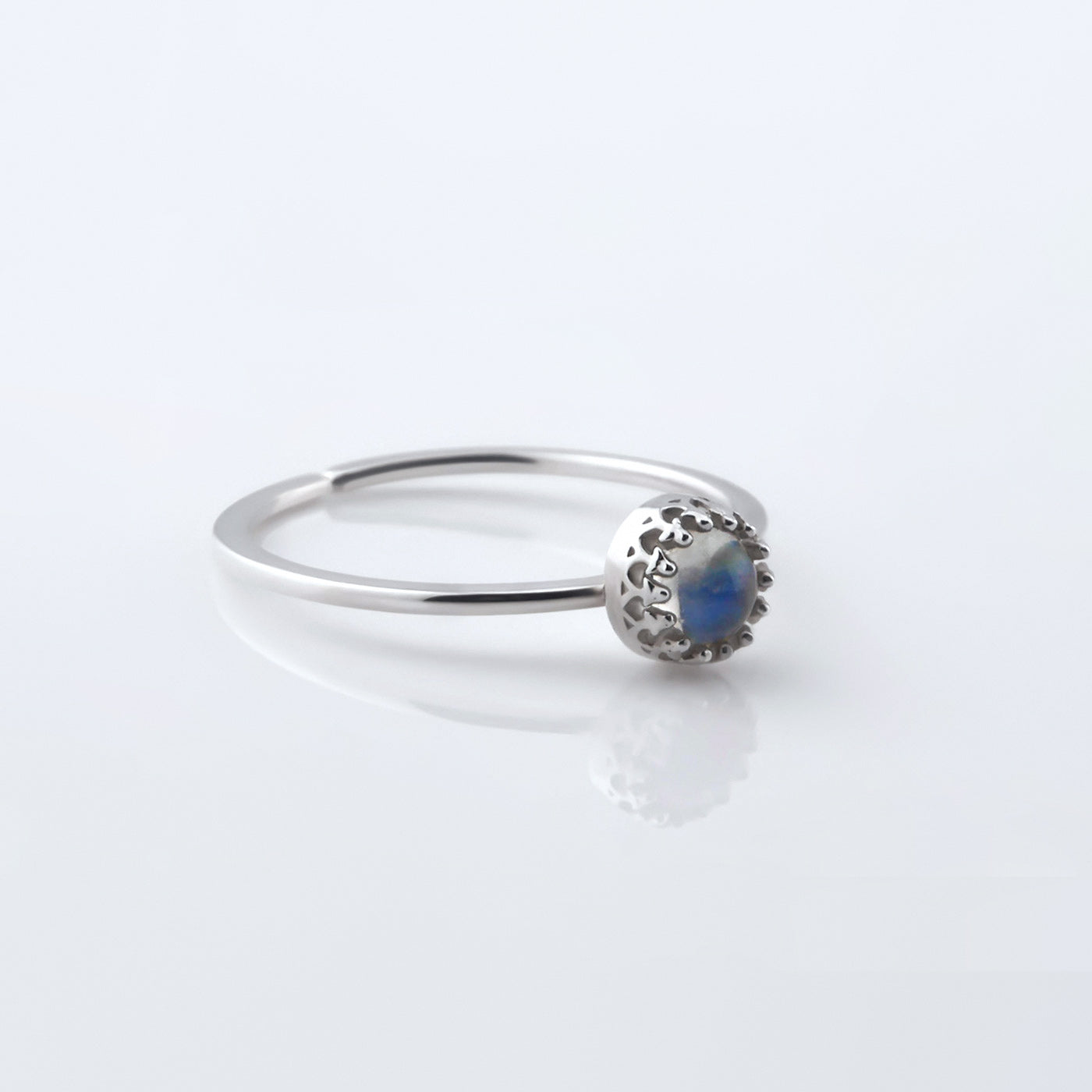 3/4 view of a dainty adjustable sterling silver ring with a focal high quality natural AAA rainbow moonstone solitaire gemstone cabochon set in a crown bezel setting. The ring is laying flat on a plain white reflective surface.