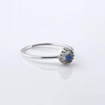 3/4 view of a dainty adjustable sterling silver ring with a focal high quality natural AAA rainbow moonstone solitaire gemstone cabochon set in a crown bezel setting. The ring is laying flat on a plain white reflective surface.