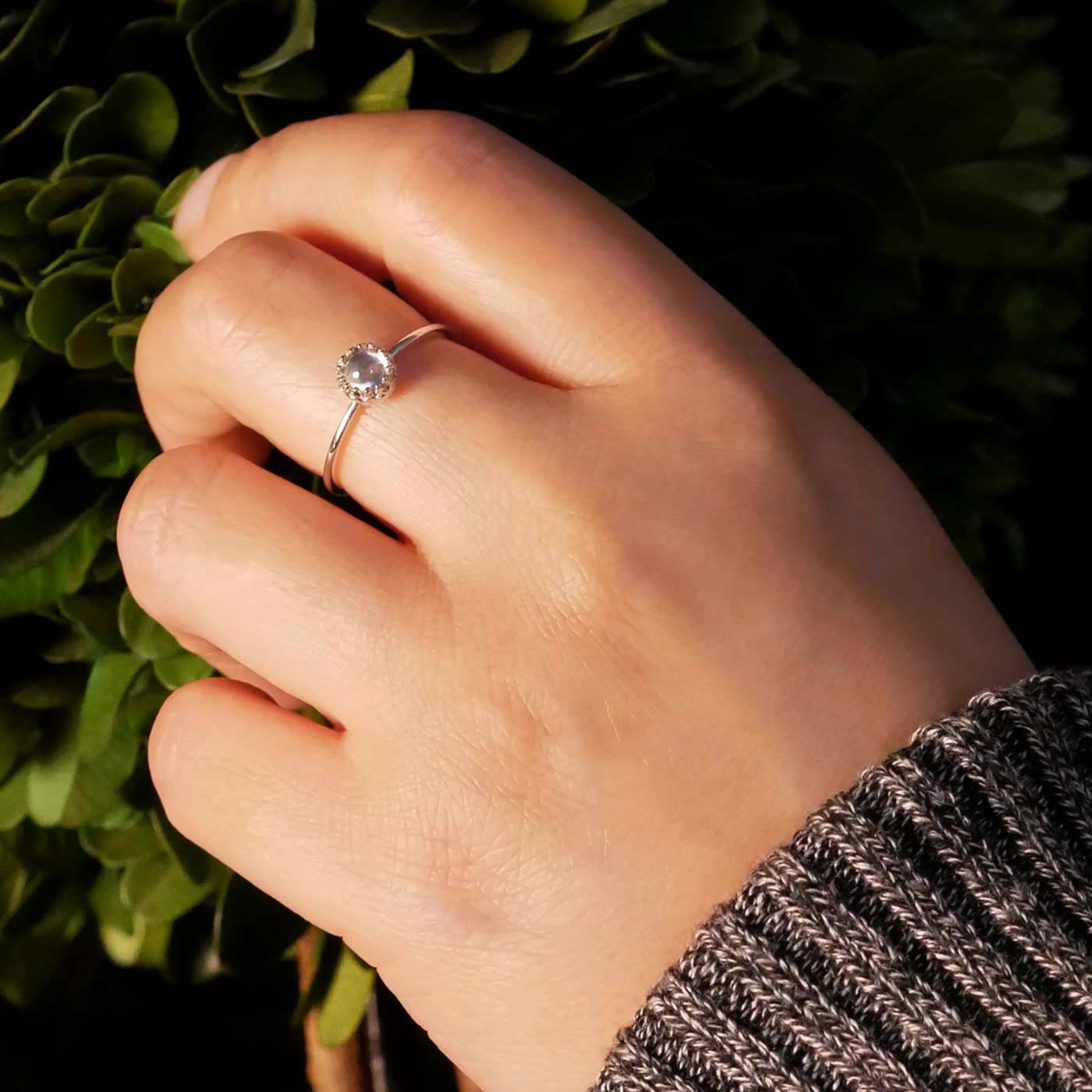 Hand wearing a dainty adjustable sterling silver ring with a focal natural high quality AAA rainbow moonstone solitaire gemstone cabochon set in a crown bezel setting. The person has a gray textured sleeve on with background of green plant leaves on a small plant.