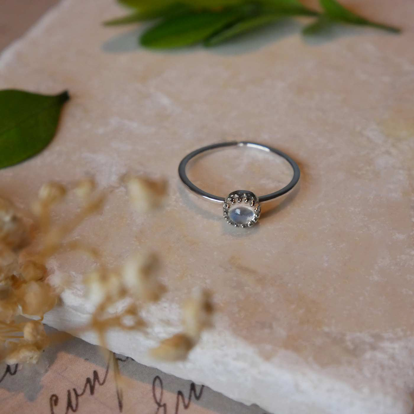 Top front view of a dainty adjustable sterling silver ring with a focal natural high quality AAA rainbow moonstone solitaire gemstone cabochon set in a crown bezel setting laying on a stone surface over textured vintage beige pink paper with cursive writing on it, also stylized with green leaves and blurred beige flowers in the foreground.