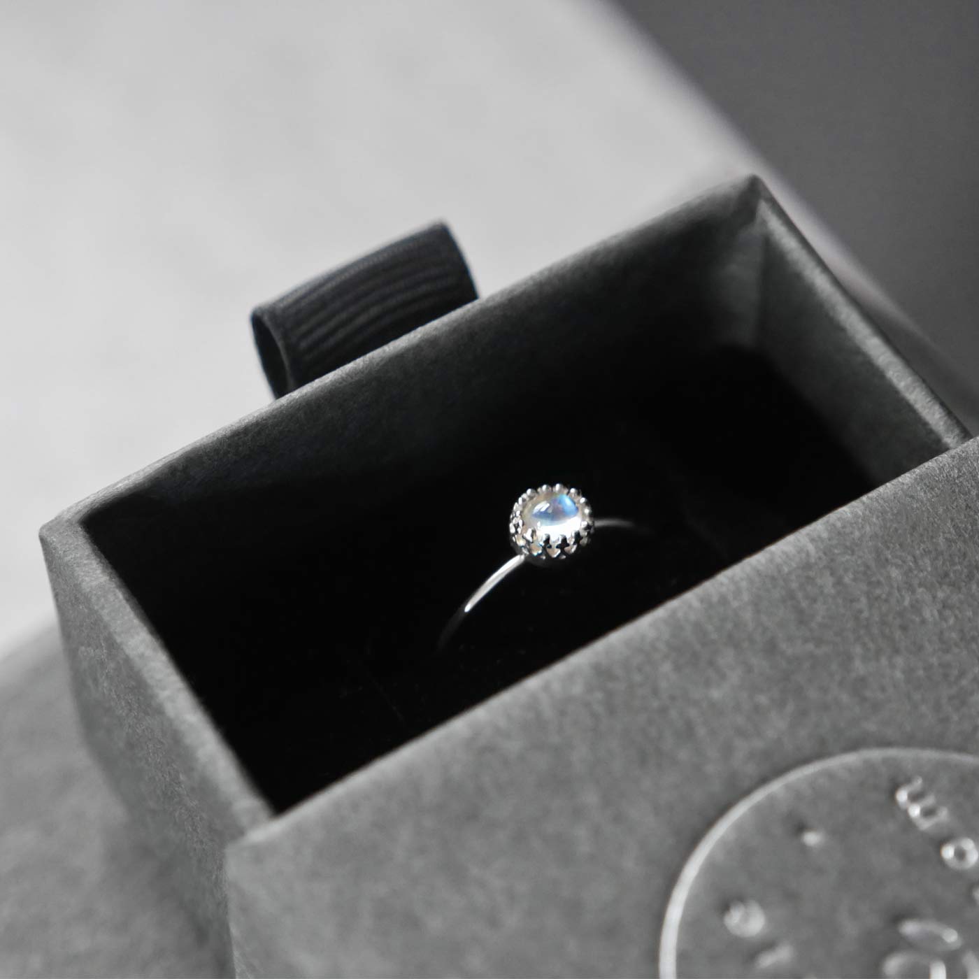 Top view of a dainty adjustable sterling silver ring with a focal natural high quality AAA rainbow moonstone solitaire gemstone cabochon set in a crown bezel setting. The ring is tucked into the black velvet insert of a gray textured colored jewelry box with reverie blossom silver brand text on it.