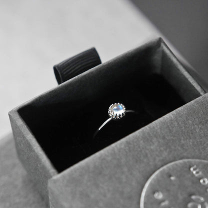 Top view of a dainty adjustable sterling silver ring with a focal natural high quality AAA rainbow moonstone solitaire gemstone cabochon set in a crown bezel setting. The ring is tucked into the black velvet insert of a gray textured colored jewelry box with reverie blossom silver brand text on it.