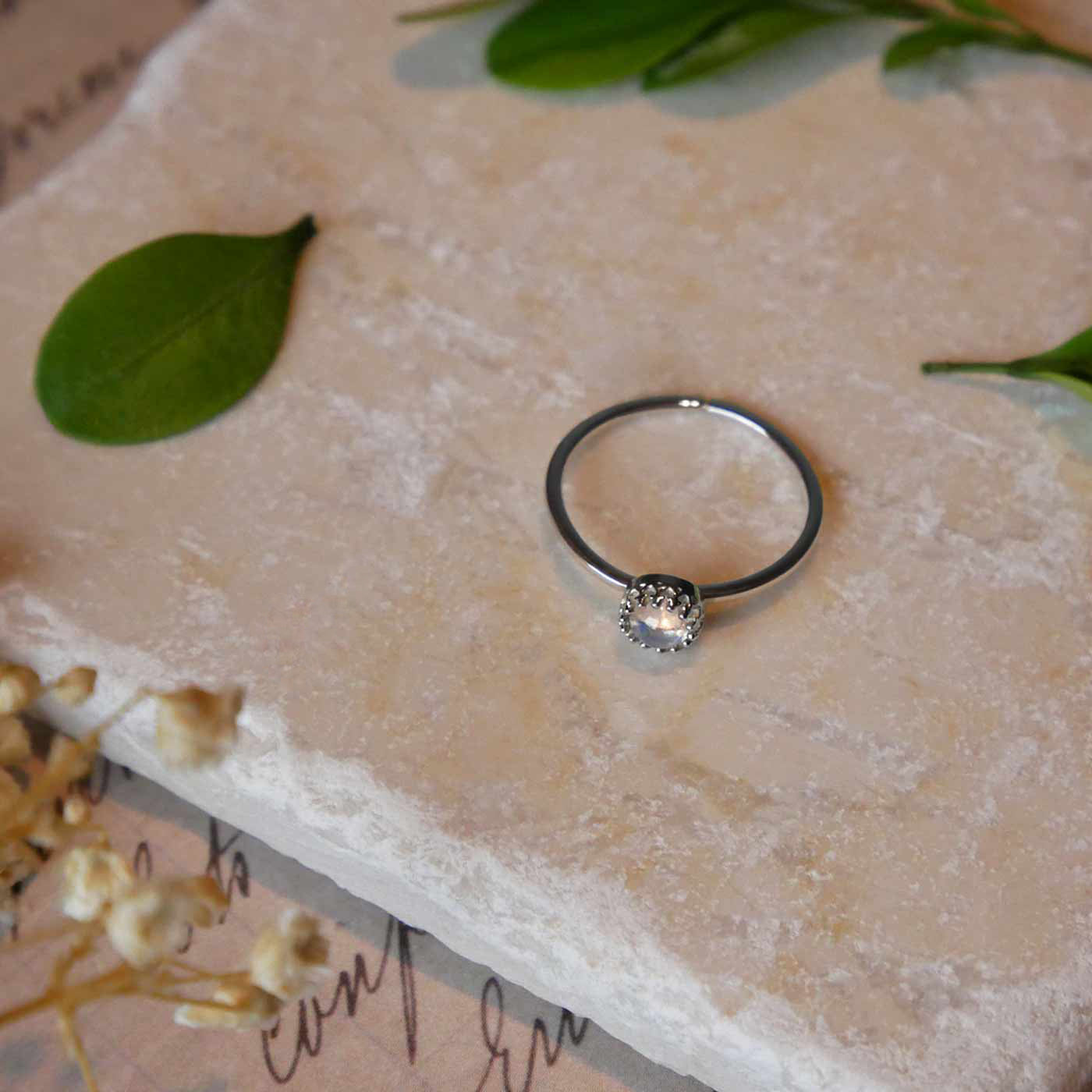 Top front view of a dainty adjustable sterling silver ring with a focal natural high quality AAA rainbow moonstone solitaire gemstone cabochon set in a crown bezel setting laying on a stone surface over textured vintage beige pink paper with cursive writing on it, also stylized with green leaves and blurred beige flowers in the foreground.