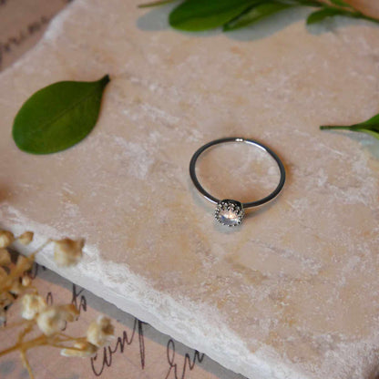 Top front view of a dainty adjustable sterling silver ring with a focal natural high quality AAA rainbow moonstone solitaire gemstone cabochon set in a crown bezel setting laying on a stone surface over textured vintage beige pink paper with cursive writing on it, also stylized with green leaves and blurred beige flowers in the foreground.