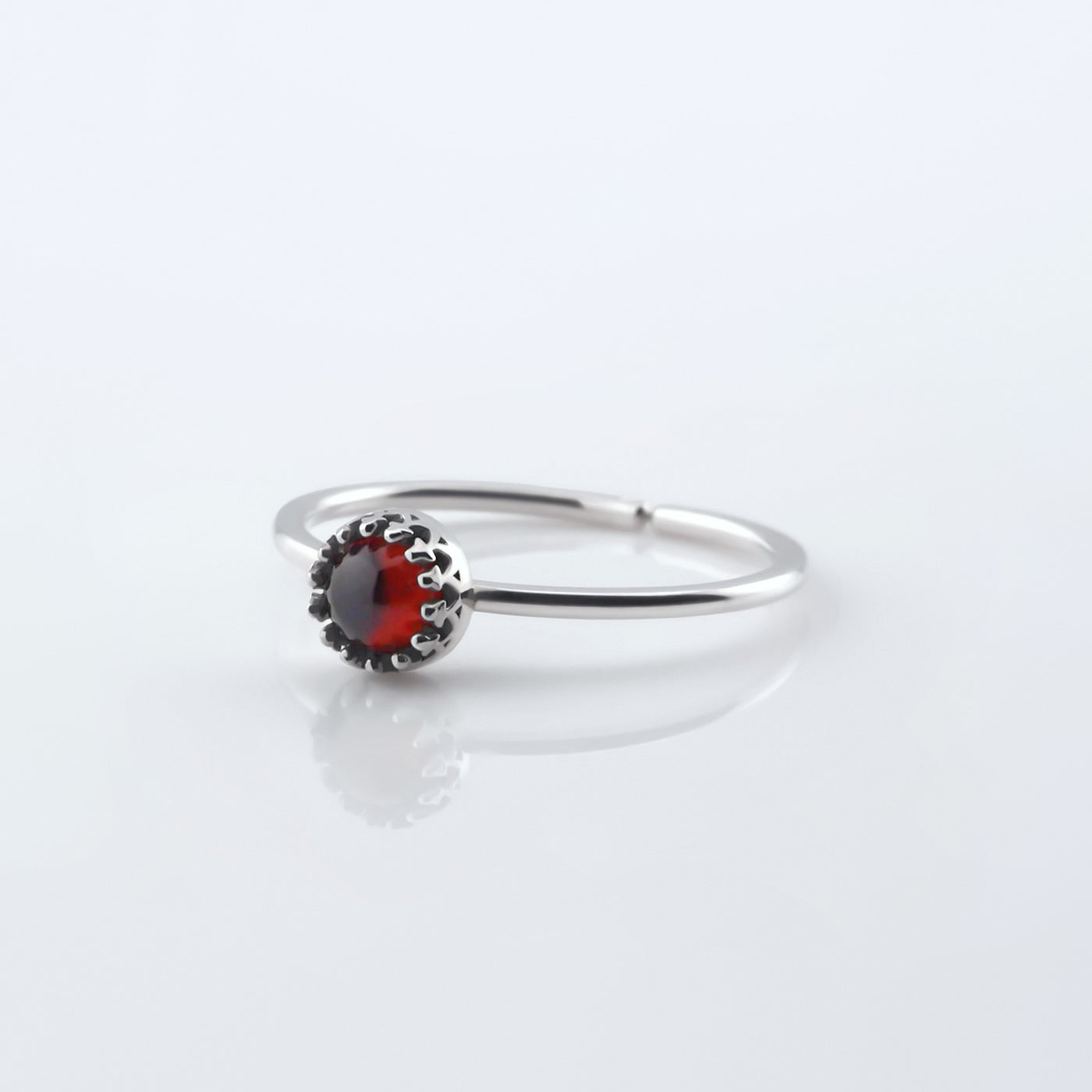 Front 3/4 view of a dainty adjustable sterling silver ring with a focal high quality natural AAA deep red hessonite garnet gemstone cabochon set in a crown bezel setting. The ring is laying flat on a plain white reflective surface.