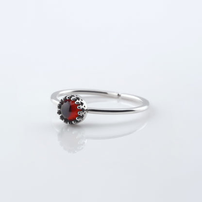 Front 3/4 view of a dainty adjustable sterling silver ring with a focal high quality natural AAA deep red hessonite garnet gemstone cabochon set in a crown bezel setting. The ring is laying flat on a plain white reflective surface.
