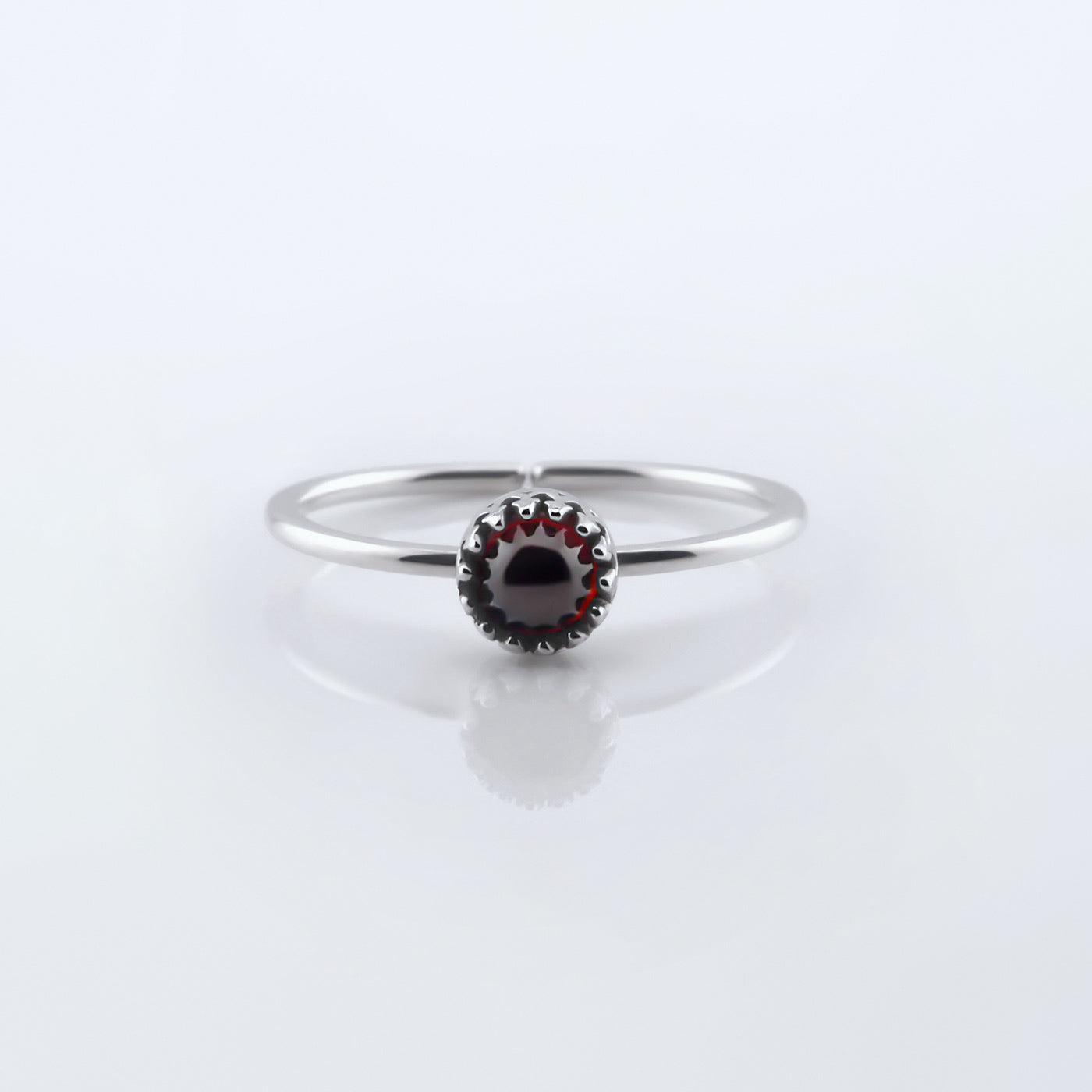 Front view of a dainty adjustable sterling silver ring with a focal high quality AAA deep red hessonite garnet cabochon set in a crown bezel setting. The ring is laying flat on a plain white reflective surface.