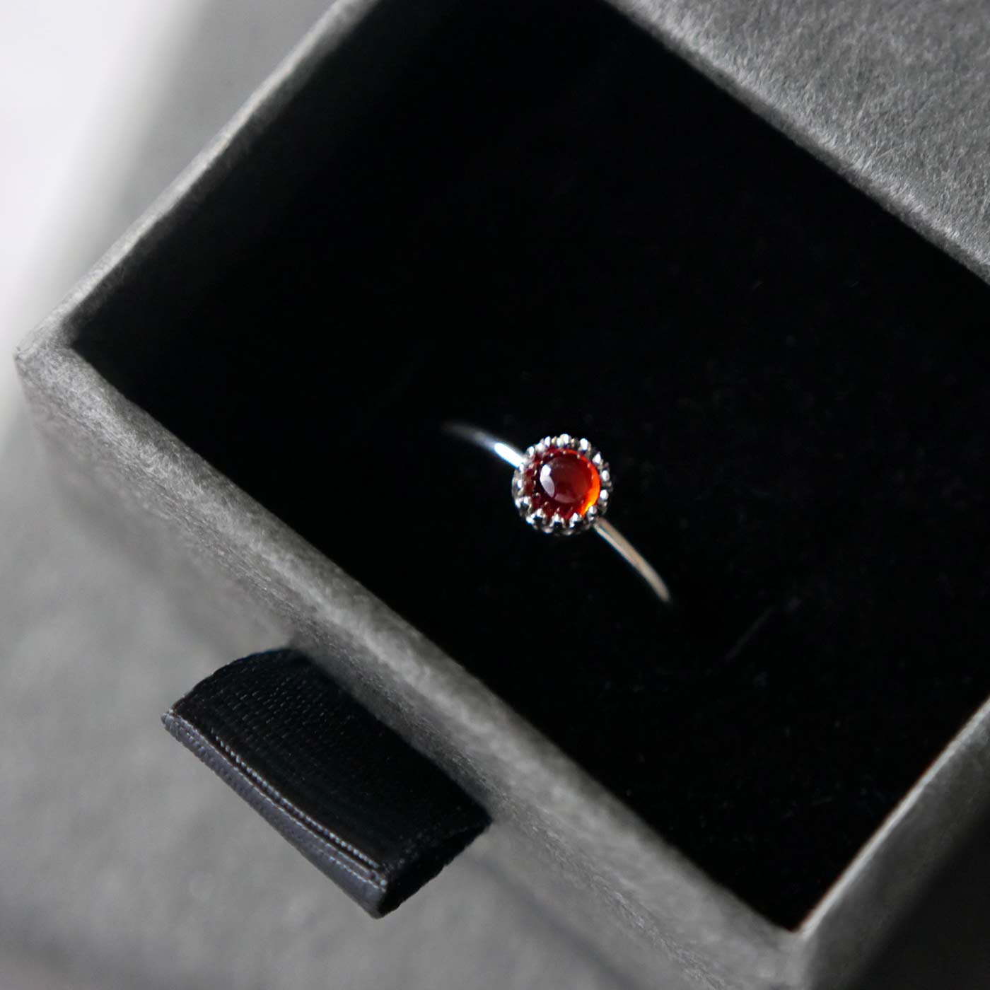Top view of a dainty adjustable sterling silver ring with a focal natural high quality aaa deep red hessonite garnet gemstone cabochon set in a crown bezel setting. The ring is tucked into the black velvet insert of a gray textured colored jewelry box.