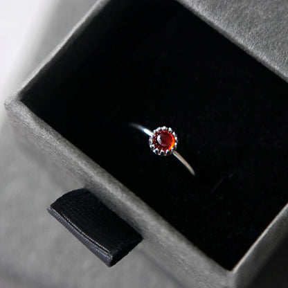 Top view of a dainty adjustable sterling silver ring with a focal natural high quality aaa deep red hessonite garnet gemstone cabochon set in a crown bezel setting. The ring is tucked into the black velvet insert of a gray textured colored jewelry box.