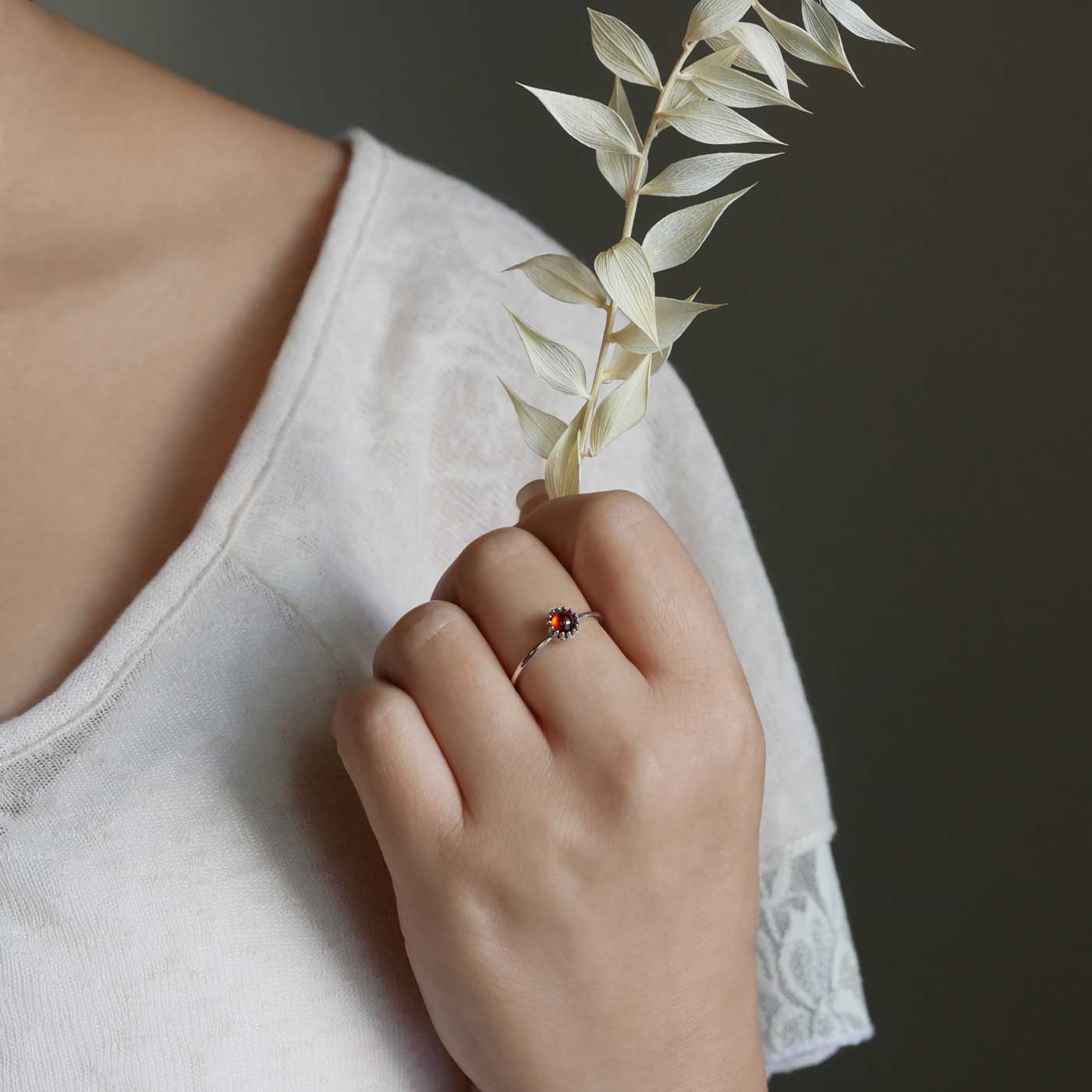 Hand holding dried baby's breath flowers with a dainty adjustable sterling silver ring with a focal high quality natural aaa deep red hessonite garnet gemstone cabochon set in a crown bezel setting. The person is wearing a beige off white colored top as the background.