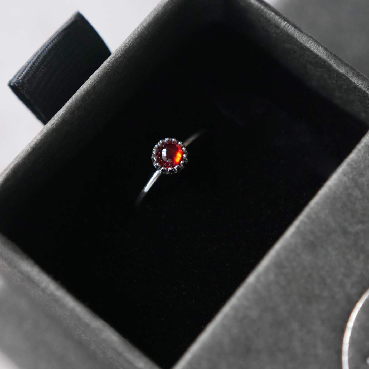 Top view of a dainty adjustable sterling silver ring with a focal natural high quality aaa deep red hessonite garnet gemstone cabochon set in a crown bezel setting. The ring is tucked into the black velvet insert of a gray textured colored jewelry box.