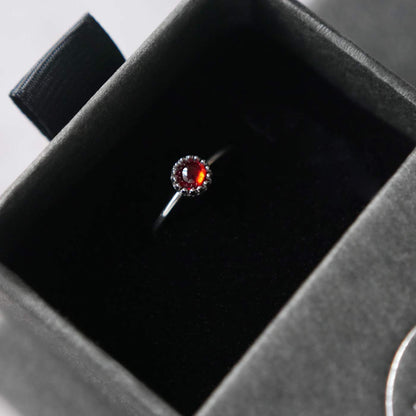 Top view of a dainty adjustable sterling silver ring with a focal natural high quality aaa deep red hessonite garnet gemstone cabochon set in a crown bezel setting. The ring is tucked into the black velvet insert of a gray textured colored jewelry box.