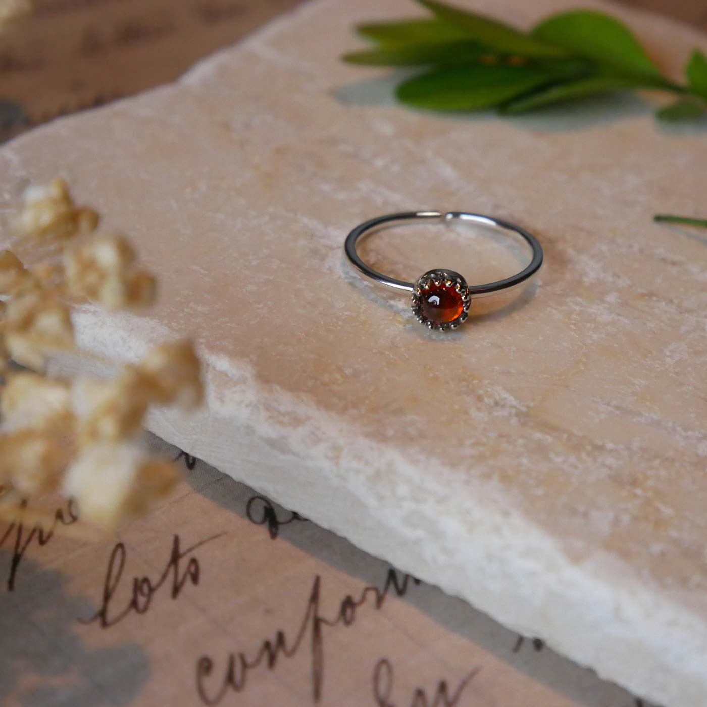 Front view of a dainty adjustable sterling silver ring with a focal natural high quality aaa deep red hessonite garnet gemstone cabochon set in a crown bezel setting laying on a stone surface over textured vintage beige pink paper with cursive writing on it, also stylized with green leaves and blurred beige flowers in the foreground.