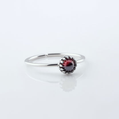 Front 3/4 view of a dainty adjustable sterling silver ring with a focal high quality aaa deep red  hessonite garnet cabochon set in a crown bezel setting. The ring is laying flat on a plain white reflective surface.