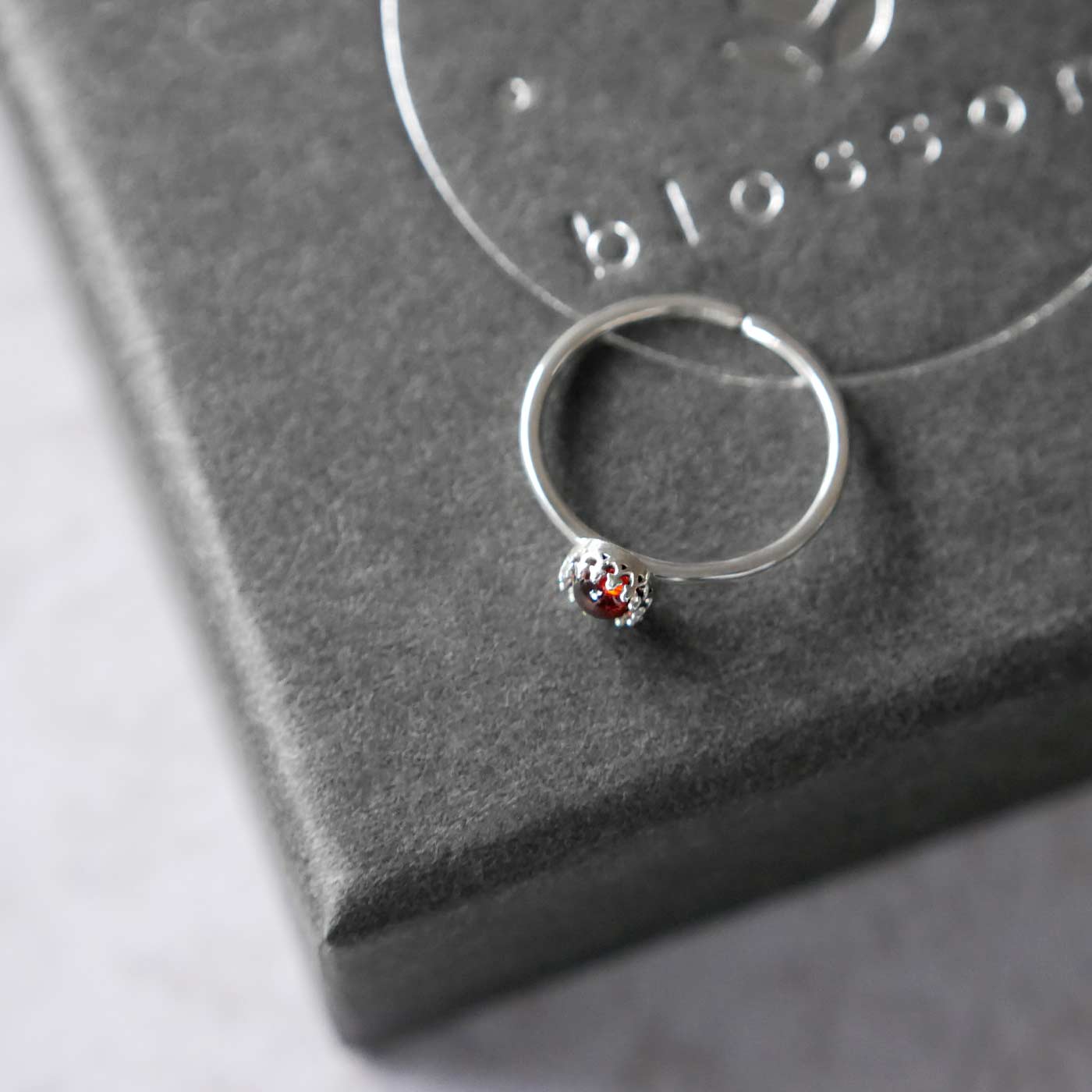 Top side view of a dainty adjustable sterling silver ring with a focal natural aaa deep red hessonite garnet gemstone cabochon set in a crown bezel setting. The ring is laying flat on a gray textured colored jewelry box with silver reverie blossom branded text.