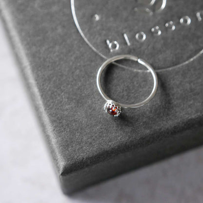 Top side view of a dainty adjustable sterling silver ring with a focal natural aaa deep red hessonite garnet gemstone cabochon set in a crown bezel setting. The ring is laying flat on a gray textured colored jewelry box with silver reverie blossom branded text.