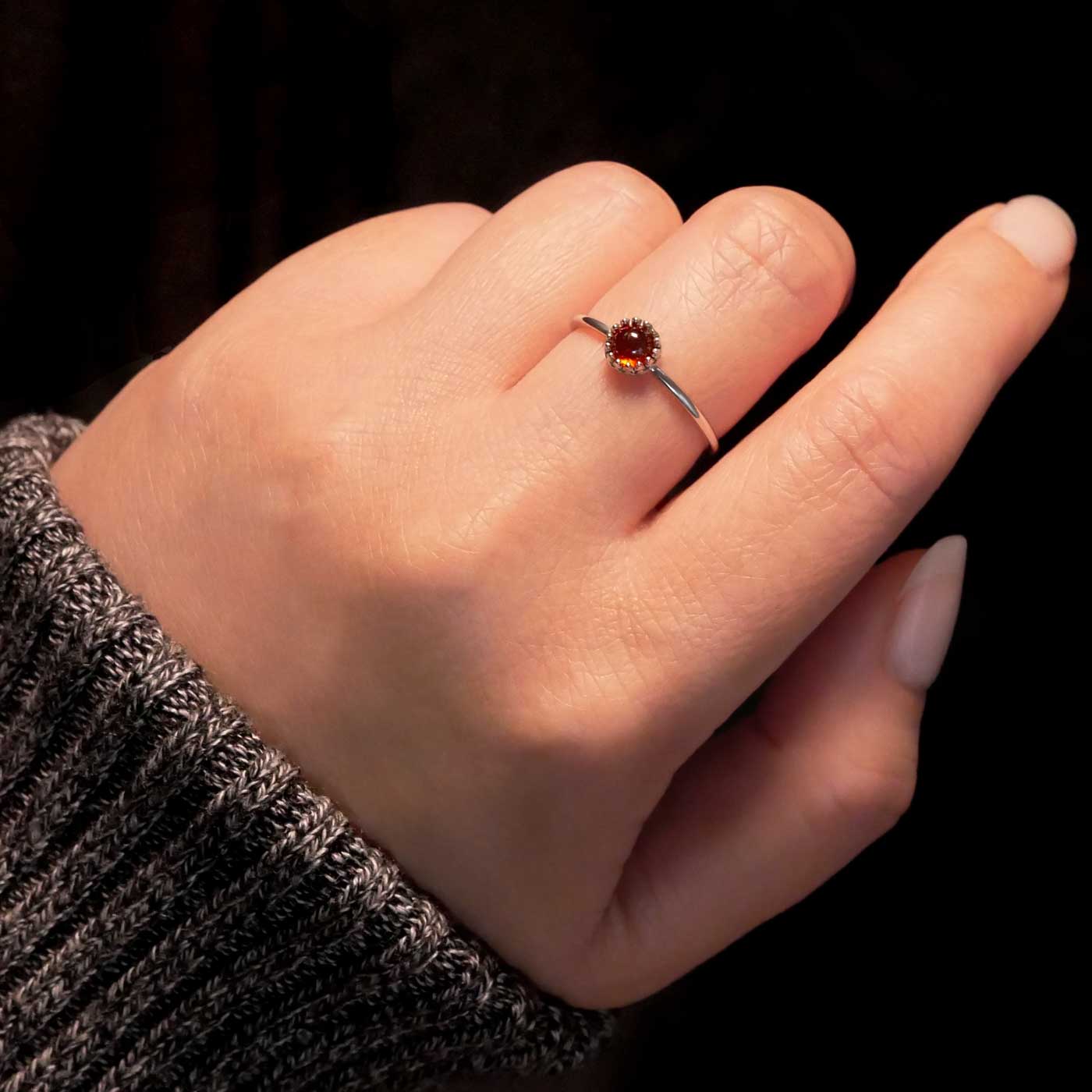 Hand wearing a dainty adjustable sterling silver ring with a focal natural high quality aaa deep red hessonite garnet gemstone cabochon set in a crown bezel setting. The person has a gray textured sleeve on with a dark black background.