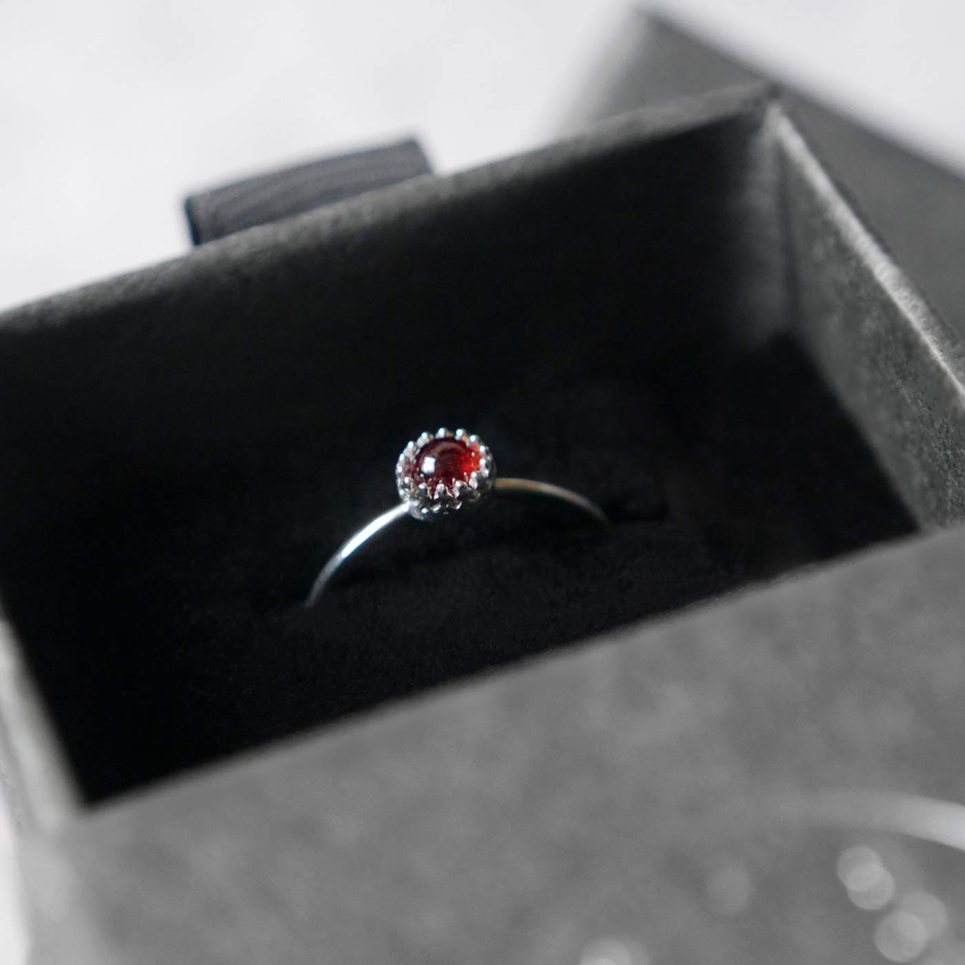 Top side view of a dainty adjustable sterling silver ring with a focal natural high quality aaa deep red hessonite garnet gemstone cabochon set in a crown bezel setting. The ring is tucked into the black velvet insert of a gray textured colored jewelry box.