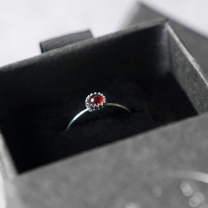 Top side view of a dainty adjustable sterling silver ring with a focal natural high quality aaa deep red hessonite garnet gemstone cabochon set in a crown bezel setting. The ring is tucked into the black velvet insert of a gray textured colored jewelry box.