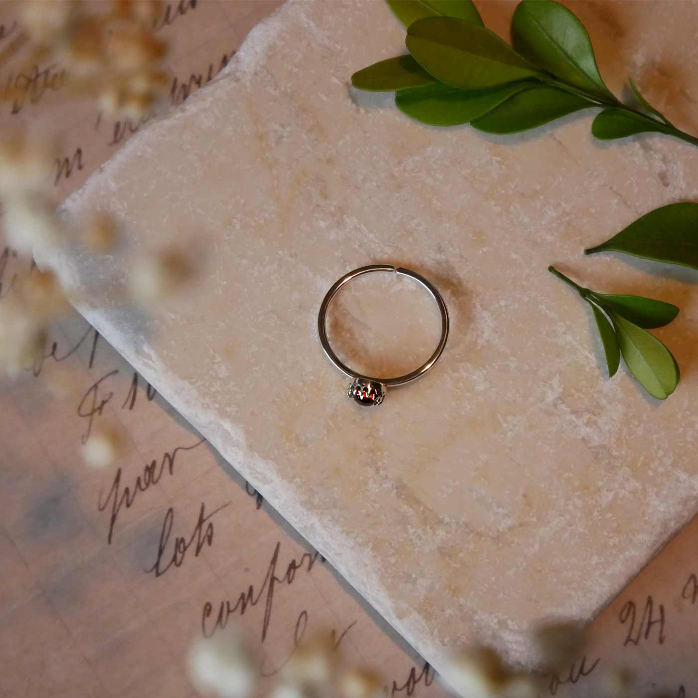 Top view of a dainty adjustable sterling silver ring with a focal natural high quality aaa deep red hessonite garnet gemstone cabochon set in a crown bezel setting laying on a stone surface over textured vintage beige pink paper with cursive writing on it, also stylized with green leaves and blurred beige flowers in the foreground.
