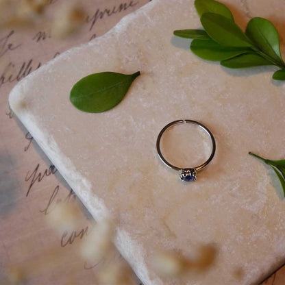 Top view of a dainty adjustable sterling silver ring with a focal natural high quality AAA blue kyanite solitaire gemstone cabochon set in a crown bezel setting laying on a stone surface over textured vintage beige pink paper with cursive writing on it, also stylized with green leaves and blurred beige flowers in the foreground.