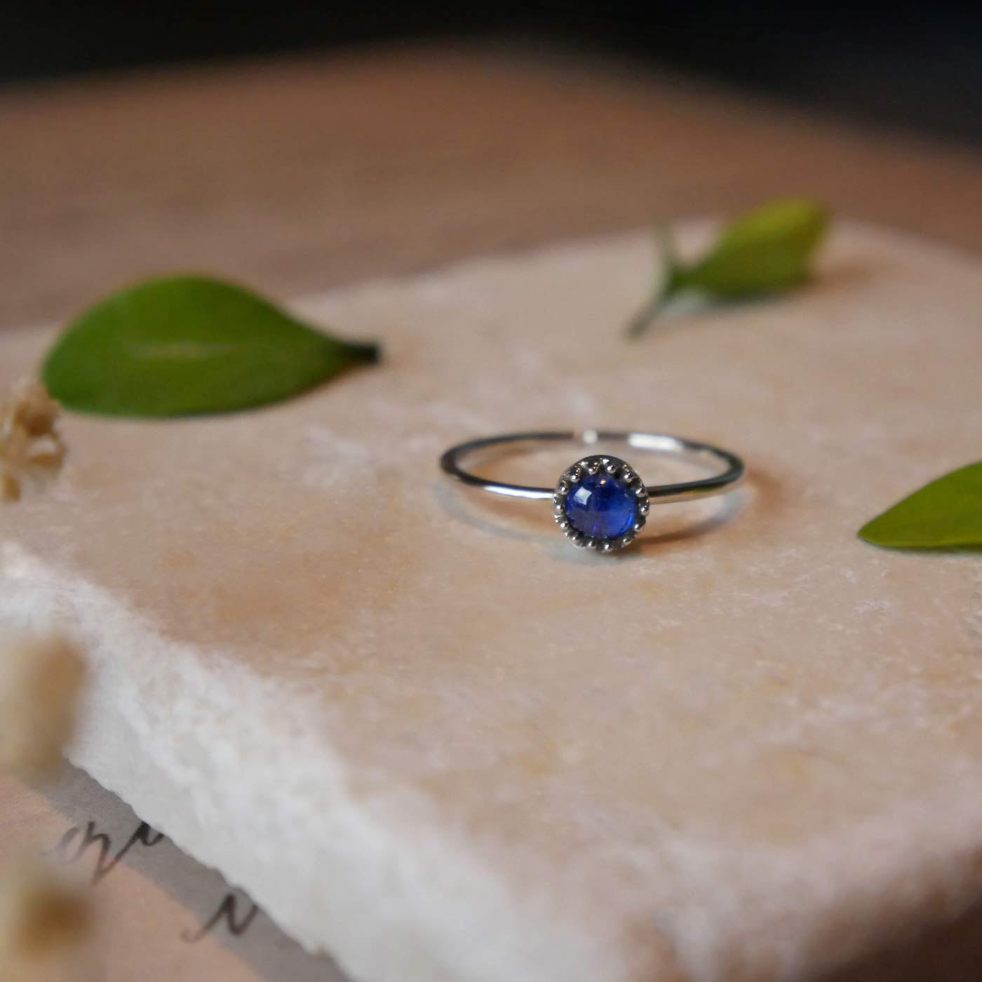 Front view of a dainty adjustable sterling silver ring with a focal natural high quality AAA blue kyanite solitaire gemstone cabochon set in a crown bezel setting laying on a stone surface over textured vintage beige pink paper with cursive writing on it, also stylized with green leaves and blurred beige flowers in the foreground.