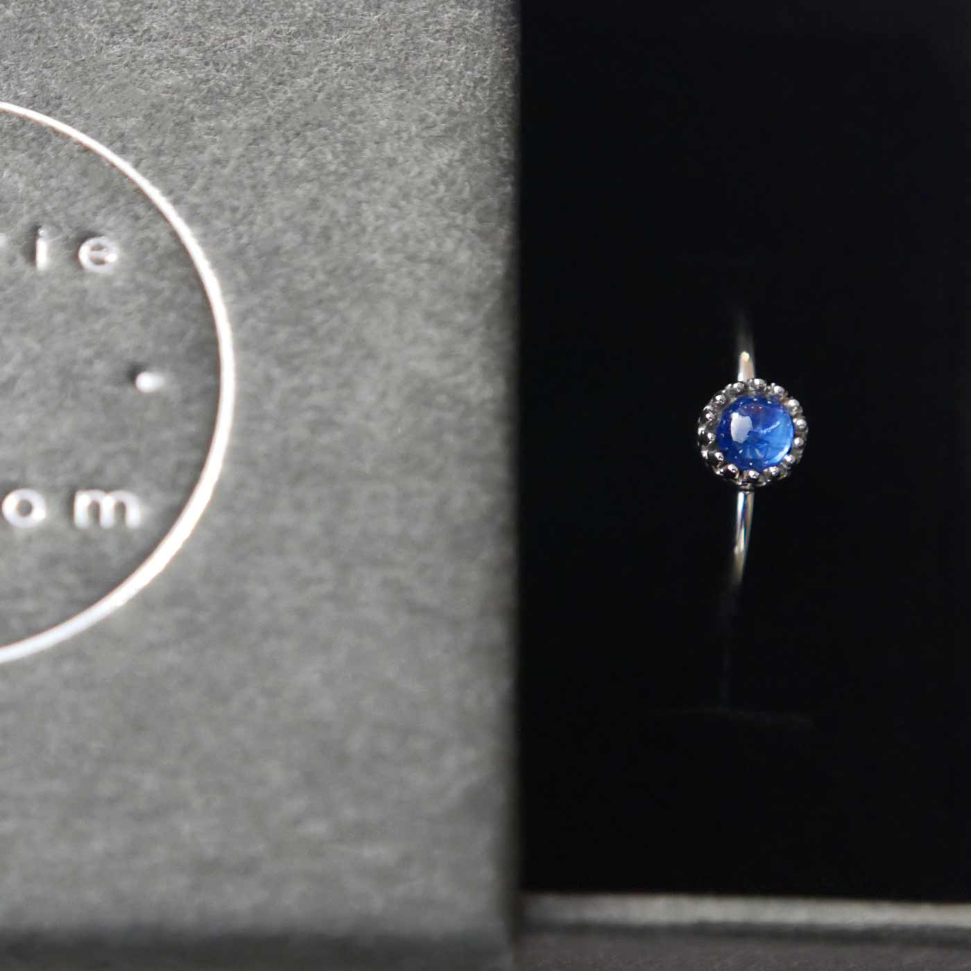 Top view of a dainty adjustable sterling silver ring with a focal natural high quality AAA blue kyanite solitaire gemstone cabochon set in a crown bezel setting. The ring is tucked into the black velvet insert of a gray textured colored jewelry box with reverie blossom silver brand text on it.