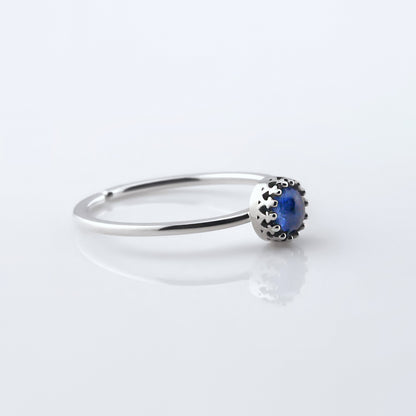 3/4 side view of a dainty adjustable sterling silver ring with a focal blue kyanite solitaire gemstone cabochon set in a crown bezel setting. The ring is laying flat on a plain white reflective surface.