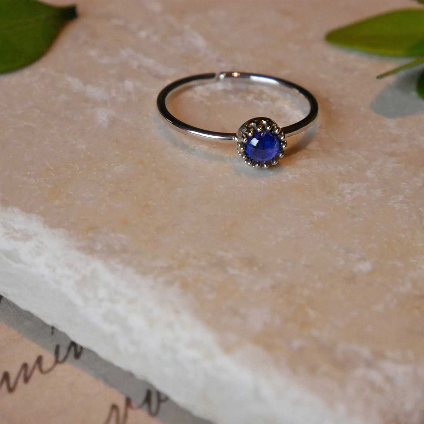 Front view of a dainty adjustable sterling silver ring with a focal natural high quality AAA blue kyanite solitaire gemstone cabochon set in a crown bezel setting laying on a stone surface over textured vintage beige pink paper with cursive writing on it, also stylized with green leaves.