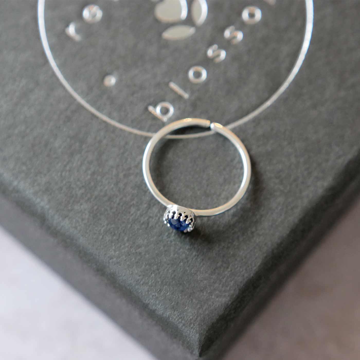 Top side view of a dainty adjustable sterling silver ring with a focal natural AAA blue kyanite solitaire gemstone cabochon set in a crown bezel setting. The ring is laying flat on a gray textured colored jewelry box with silver reverie blossom branded text.