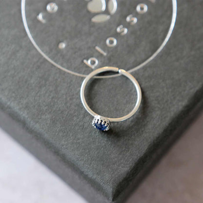 Top side view of a dainty adjustable sterling silver ring with a focal natural AAA blue kyanite solitaire gemstone cabochon set in a crown bezel setting. The ring is laying flat on a gray textured colored jewelry box with silver reverie blossom branded text.