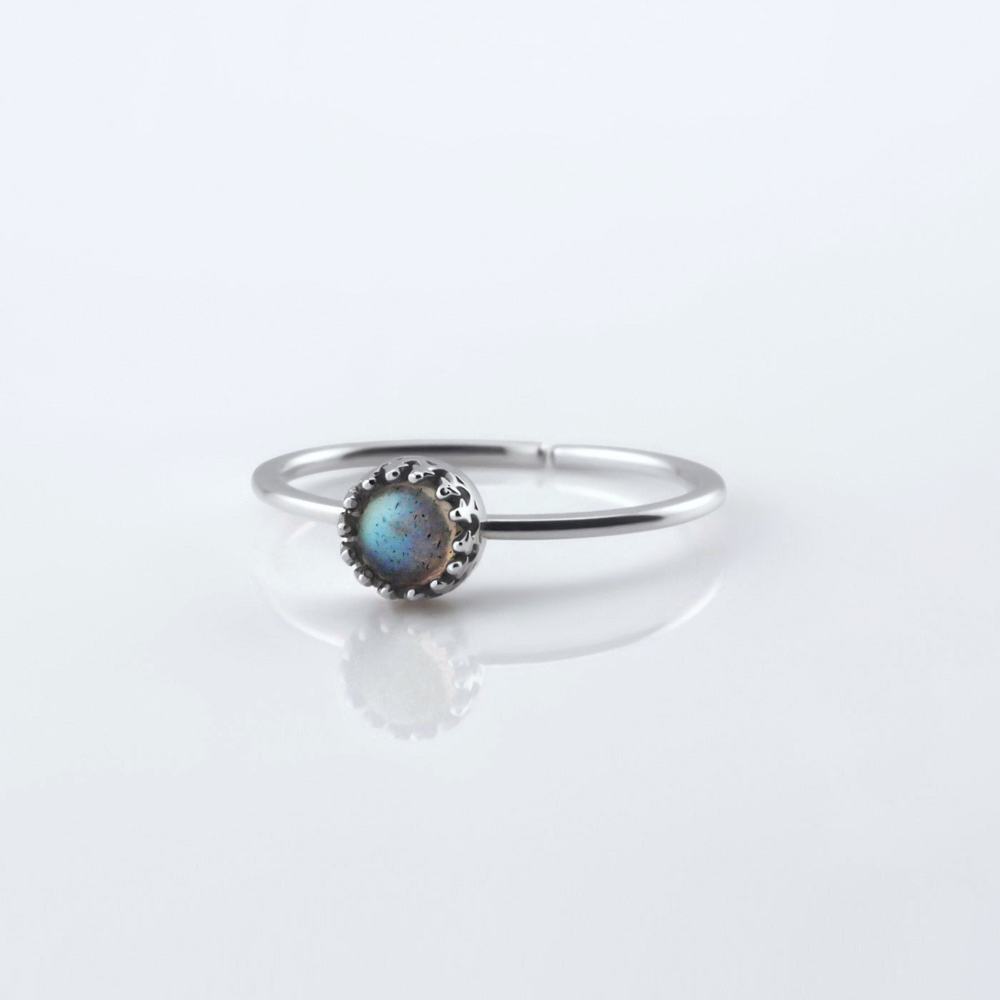 Front view of a dainty adjustable sterling silver ring with a focal labradorite gemstone cabochon set in a crown bezel setting. The ring is laying flat on a plain white reflective surface.