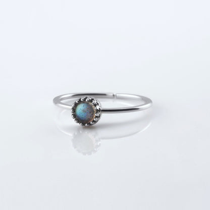 Front view of a dainty adjustable sterling silver ring with a focal labradorite gemstone cabochon set in a crown bezel setting. The ring is laying flat on a plain white reflective surface.