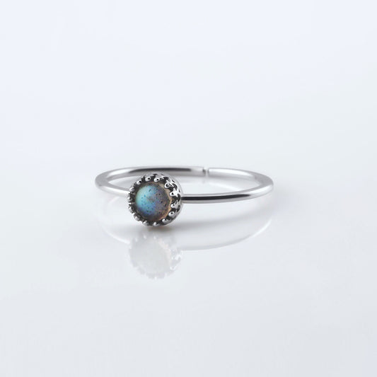 Front view of a dainty adjustable sterling silver ring with a focal labradorite gemstone cabochon set in a crown bezel setting. The ring is laying flat on a plain white reflective surface.