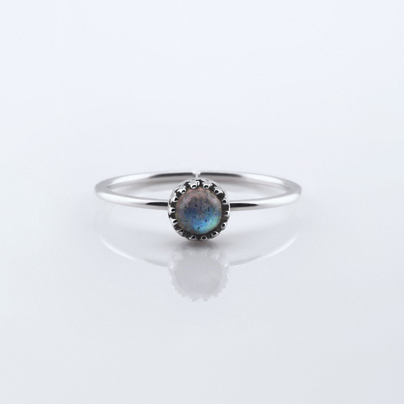 Front view of a dainty adjustable sterling silver ring with a focal high quality AAA labradorite cabochon set in a crown bezel setting. The ring is laying flat on a plain white reflective surface.