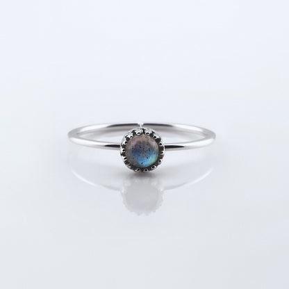 Front view of a dainty adjustable sterling silver ring with a focal high quality AAA labradorite cabochon set in a crown bezel setting. The ring is laying flat on a plain white reflective surface.
