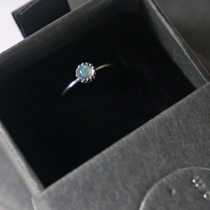 Top view of a dainty adjustable sterling silver ring with a focal natural high quality aaa labradorite gemstone cabochon set in a crown bezel setting. The ring is tucked into the black velvet insert of a gray textured colored jewelry box.